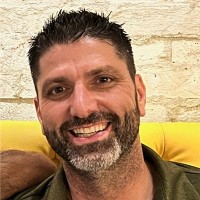 delray business partner member edmund schlacher - dark haired, brown-eyed man with greying beard, green shirt, and smile sitting on yellow couch